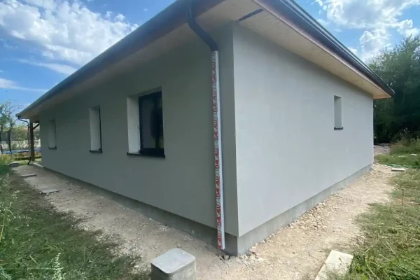 Vue extérieure d'une maison moderne avec une finition en crépi gris. La maison est de plain-pied avec un toit plat et des gouttières rouges. La façade présente un design minimaliste avec peu de fenêtres.