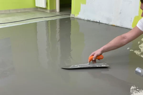 Application d'un revêtement de sol en béton ciré gris avec une spatule par une personne dont seul le bras est visible. La surface est lisse et réfléchissante, avec des reflets de la pièce environnante.